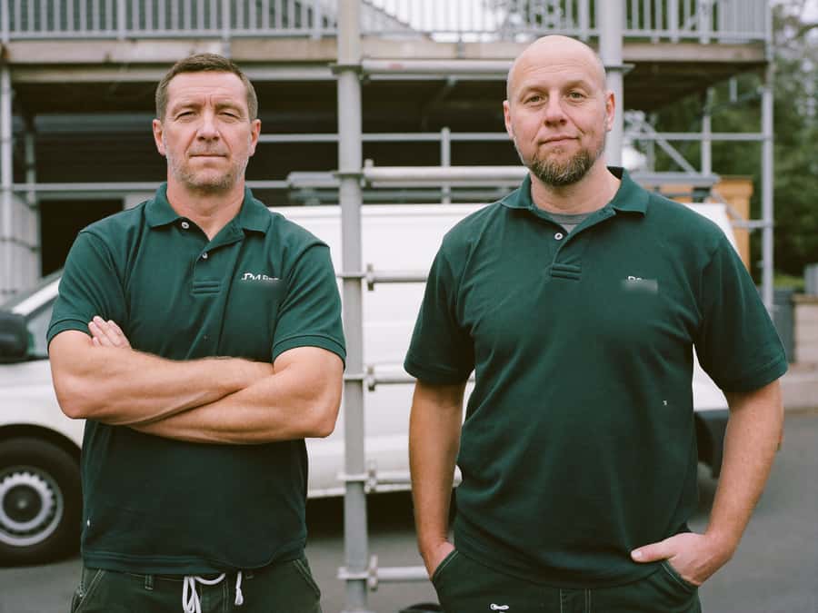Martin and Tom Halstead in matching green work shirts in front of a scaffold tower, with the firm's van behind them